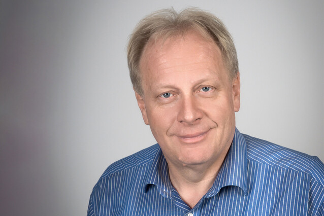 A middle-aged blond man with a blue shirt smiles at the camera.