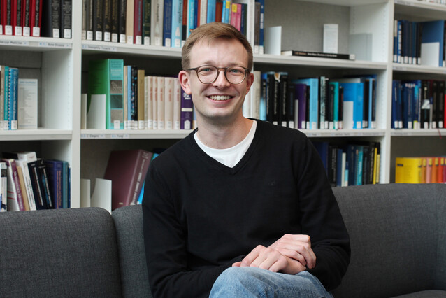 A young man with blond hair and glasses sits on a grey couch with crossed legs and arms, smiling at the camera.