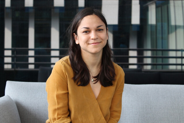 A young woman wearing a mustard yellow shirt sits on a pale grey couch. She has long dark hair and a faint smile.