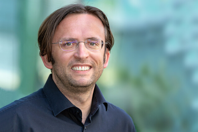 A middle-aged man with brown hair, glasses, and a black shirt smiles at the camera.