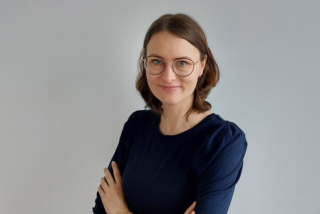 A young woman with brown hair and blue eyes, wearing glasses and a dark blue top, smiles at the camera with her arms crossed.