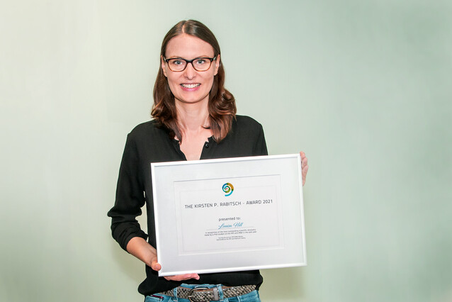 A young woman with a black shirt faces the camera and holds a silver frame that contains the award.