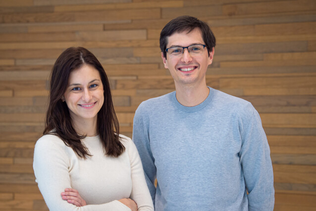 A woman on the left and a man on the right look and smile at the camera. Wooden background.