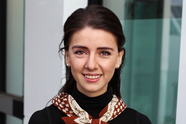 Portrait of a smiling young woman with tied, long, dark hair. She is wearing a black turtleneck and a colourful scarf.