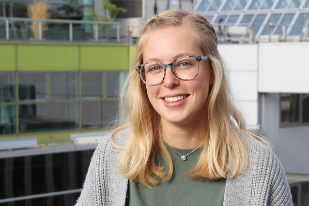 a young blond woman with glasses smiles at the camera. She's wearing a grey jacket and a green shirt.