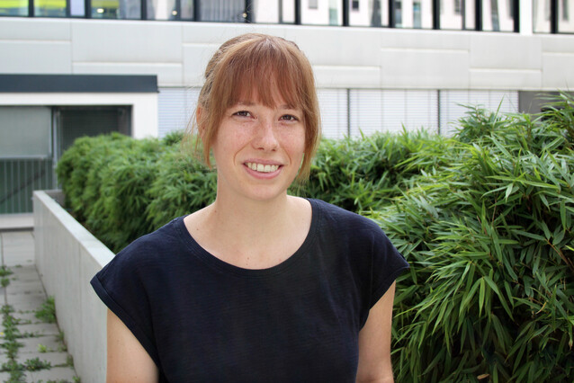 A young woman with red hair and a dark shirt smiles at the camera, with bamboo bushes in the background.