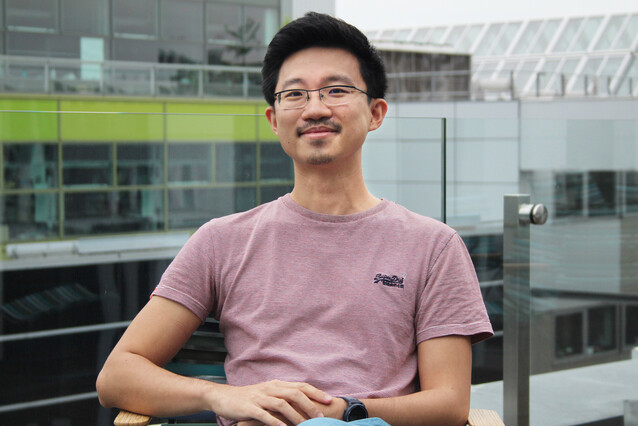 A young Asian man with a reddish t-shirt and glasses sits on a terrace and smiles at the camera.