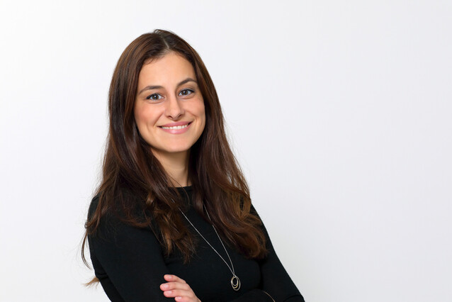 A young woman with long brown hair and a black weater smiles at the camera, with arms crossed.