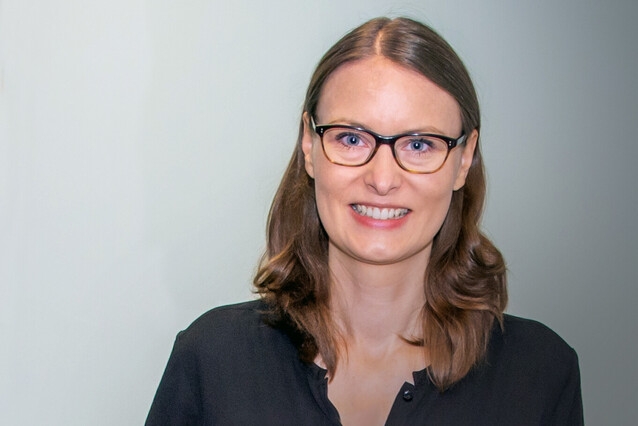 A young woman wearing glasses smiles at the camera. She has long brown hair and a black shirt.