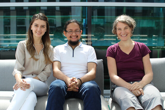 Three people sit on a couch in the sun, on the left a young woman with a beige shirt and white pants, in the middle a young bearded man with a white t-shirt and jeans, and on the right a woman with a dark red t-shirt and grey pants.