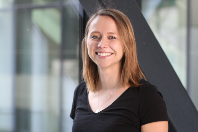 A young woman with blond hair and blue eyes smiles at the camera. She is wearing a black v-neck t-shirt.