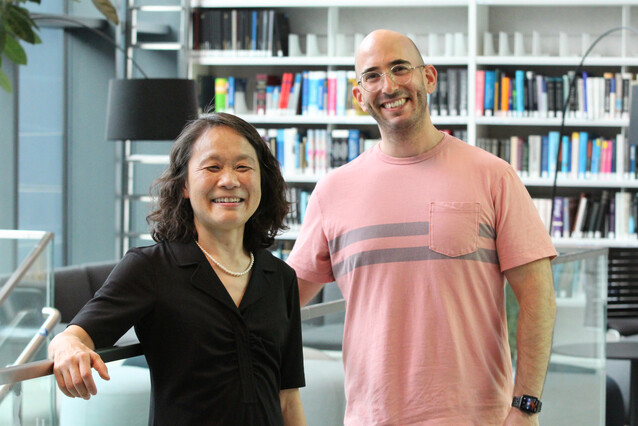 A middle-aged Asian woman with a black dress stands on the left, a young white man with glasses stands on the right.