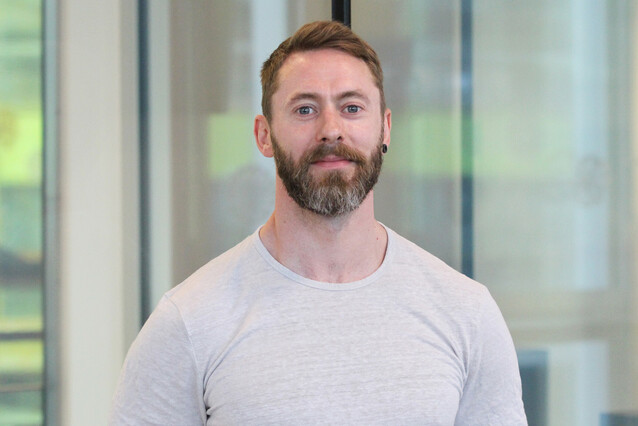 A young man with blond hair, a beard, and blue eyes and a white t-shirt smiles at the camera. 