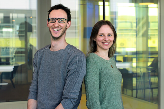 Two young people, a man (left) and a woman (right) stand back to back and smile at the camera.