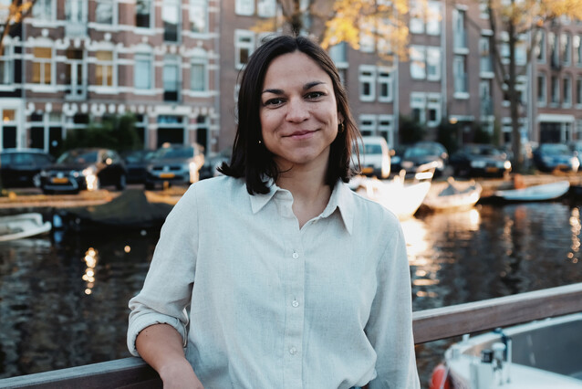 A young woman with short dark hair leans on a metallic fence and faces the camera with a smile. Behind her, a canal and red-brick houses typical of the Netherlands.