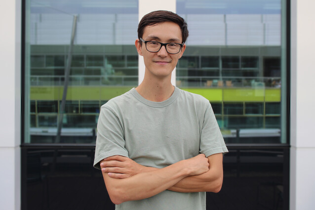 A young man with dark short hair, dark glasses, and a grey-green t-shirt crosses his arms and looks at the camera.