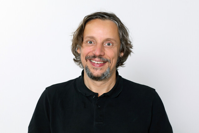 A middle-aged man with a grey beard, brown hair and pale eyes smiles at the camera.