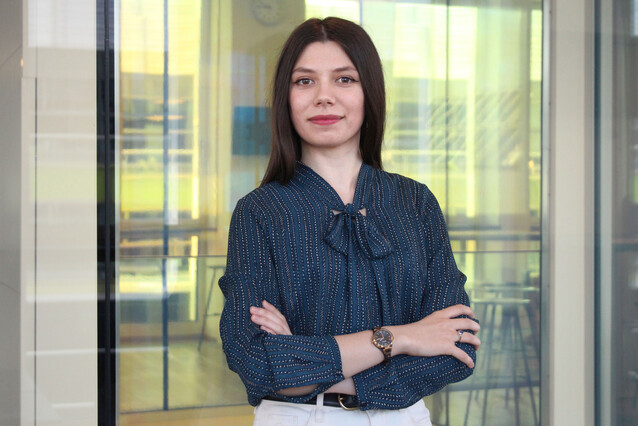 A young woman with straight brown hair looks at the camera, cross-armed. She wears a dark blue top and light pants, in front of a glass surface.