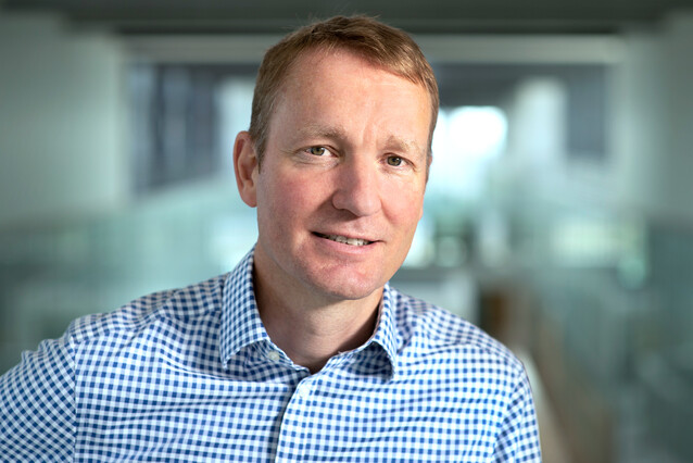 A middle-aged man with reddish hair and a checkered blue shirt smiles at the camera.