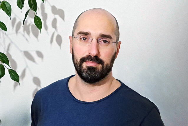 A man with a dark beard, no hair and glasses is looking at the camera with a faint smile.