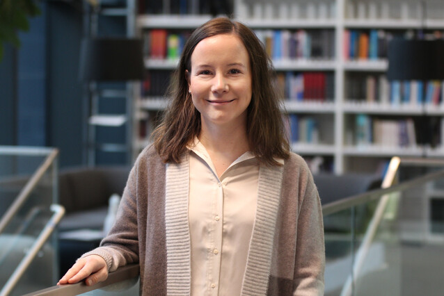 A young white woman with dark brown hair stands and looks at the camera. 